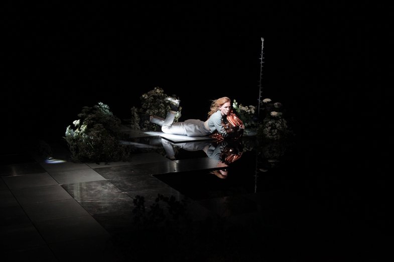 A white person is lying on the floor. Several bouquets of flowers can be seen around them and the person is illuminated by a spotlight.