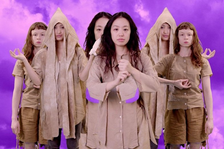 Several people stand staggered one behind the other. They are all wearing beige clothes. The background is purple.