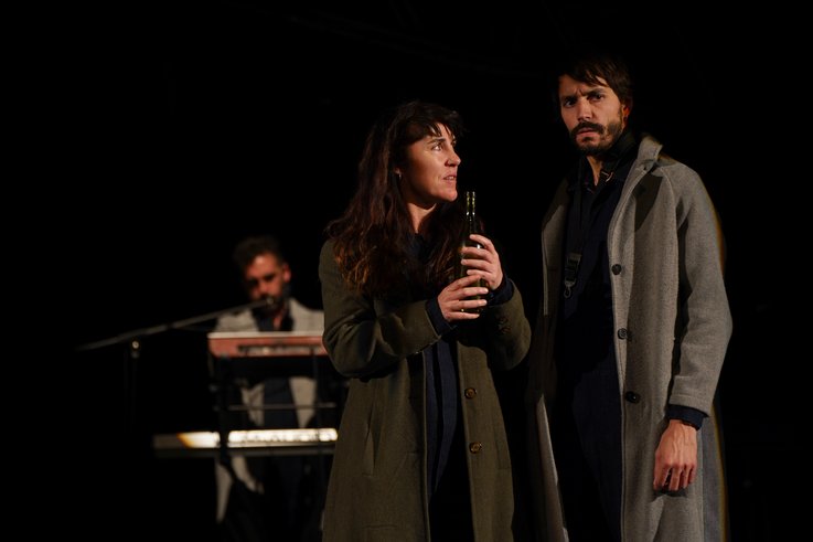 Ein Mann und eine Frau in Wintermänteln stehen vorne. Die Frau hält eine Glasflasche in der Hand. Im Hintergrund steht ein Musiker.