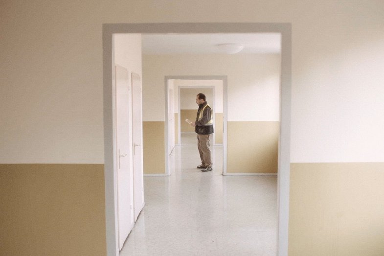 A person dressed in light brown stands in an empty hallway.