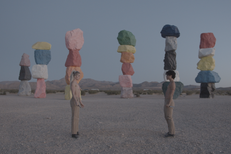 The two people face each other shirtless on a square in the mountains. In the background are colored boulders stacked on top of each other in a row.