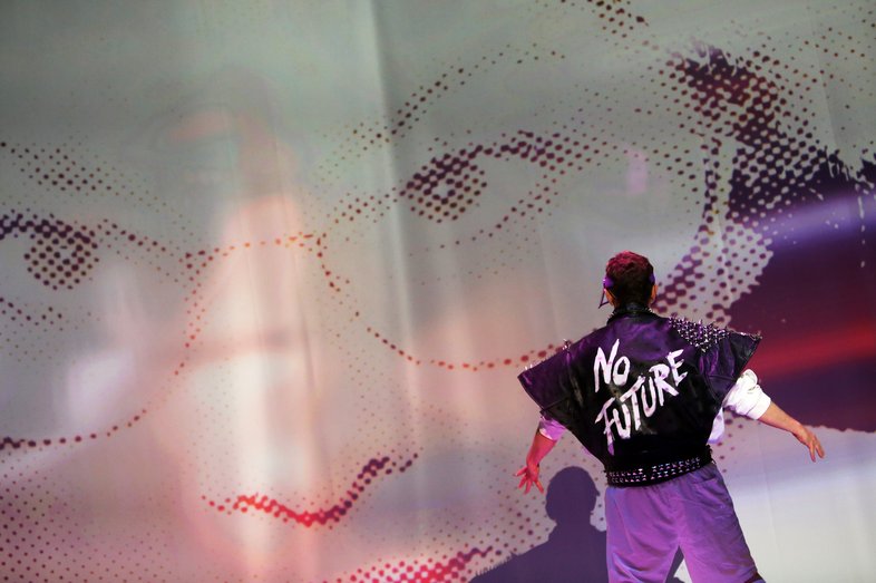 A person stands with their back to the camera, wearing a black leather jacket with "No Future" written on it. In the background, probably projected onto the stage in large format, is a pixelated image of a person wearing glasses.