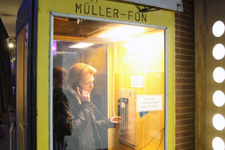 A red-haired person in a leather jacket stands in a yellow telephone booth with "Müller-Fon" written on the outside.
