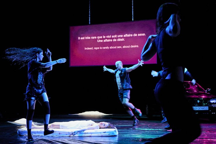 A Stage scene in which Carolina Bianchi lies unconscious on a mattress. Three people dance and jump around her. A text is projected in the background: Indeed, rape is rarely about sex, about desire.