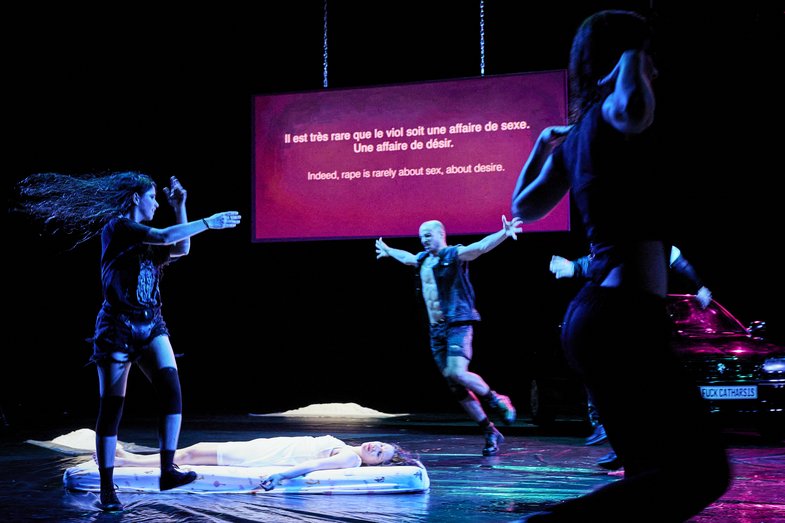 A Stage scene in which Carolina Bianchi lies unconscious on a mattress. Three people dance and jump around her. A text is projected in the background: Indeed, rape is rarely about sex, about desire.