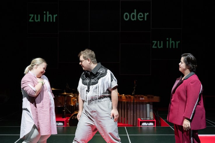 Three people on stage in a row. The person in the middle is torn between the back and forth. The words "zu ihr oder zu ihr" ("to her or to her") are projected in the background.