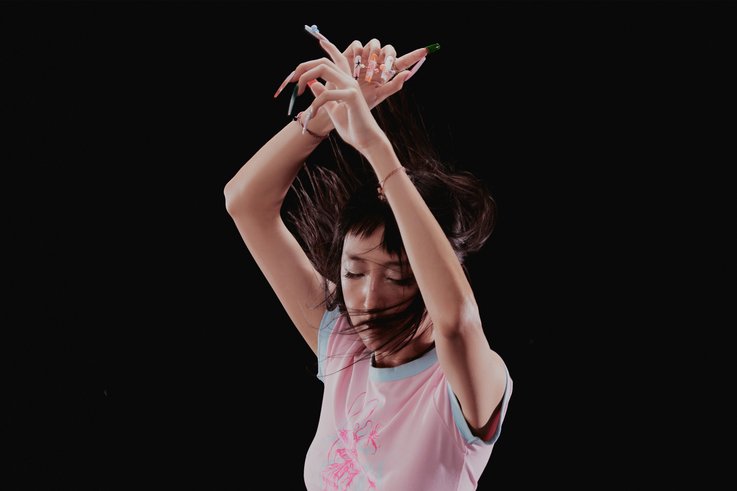 Chiro, eine DJ, hebt die Arme über den Kopf und verschränkt die Hände. Sie trägt ein helles T-Shirt mit einem Muster in der Mitte. Der Hintergrund ist schwarz.