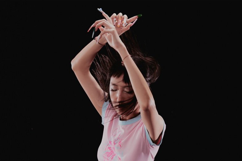 Chiro, eine DJ, hebt die Arme über den Kopf und verschränkt die Hände. Sie trägt ein helles T-Shirt mit einem Muster in der Mitte. Der Hintergrund ist schwarz.