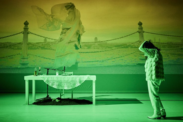 Photography of a stage scene. In the centre is a table. On the left in the foreground is a person in a dismissive posture. In the background is a projected scene of a veiled person on a coastal promenade.