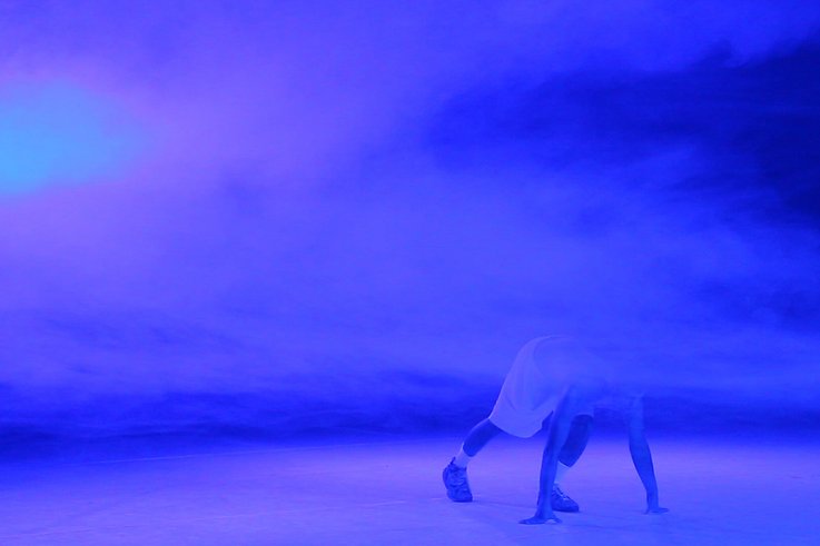 In der Mitte des Bildes steht eine Person mit ausgestreckten Armen. Die Bühne ist in blaues Licht und Nebel gehüllt.