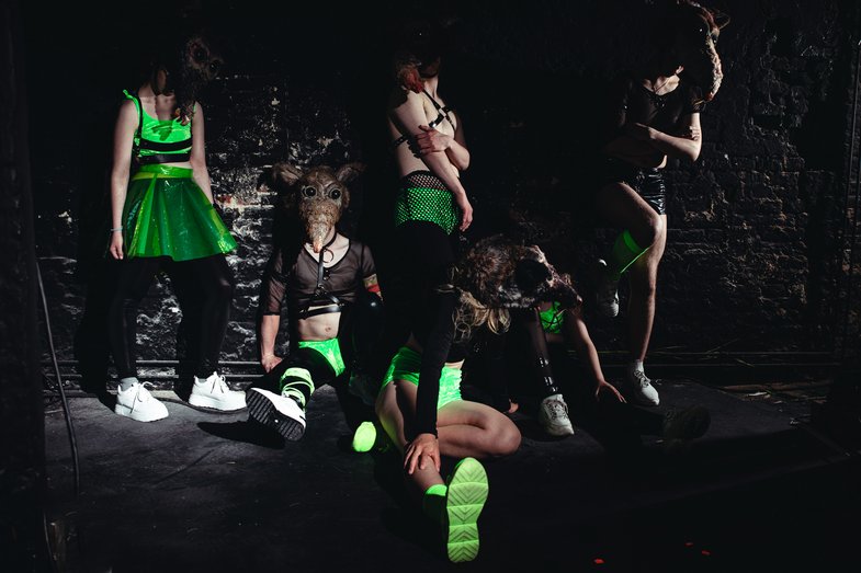 Six people wearing animal-like masks stand and sit in a black-painted room.