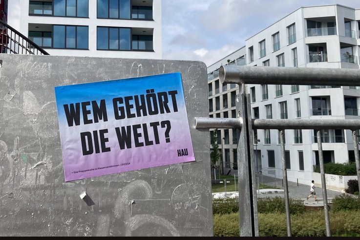 Fotografie aus dem urbanen Raum. Im Vordergrund ist ein Plakat des HAU abgebildet. Darauf steht: “Wem gehört die Welt?“