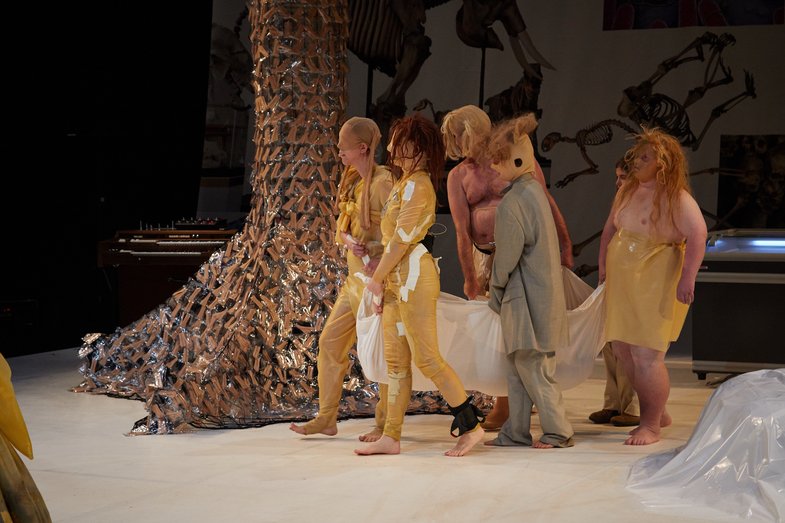 Sechs Personen im Kostüm tragen ein weißes Laken, in dem sich eine weitere Person befindet.  Sie gehen durch das Bühnenbild. Im Hintergrund befindet sich ein abstrakter Baum.