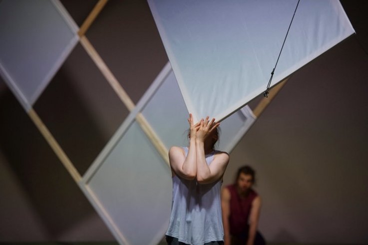 A person balances a wooden frame covered with linen in front of his face. In the background is another wooden frame on which square linen is partially covered.