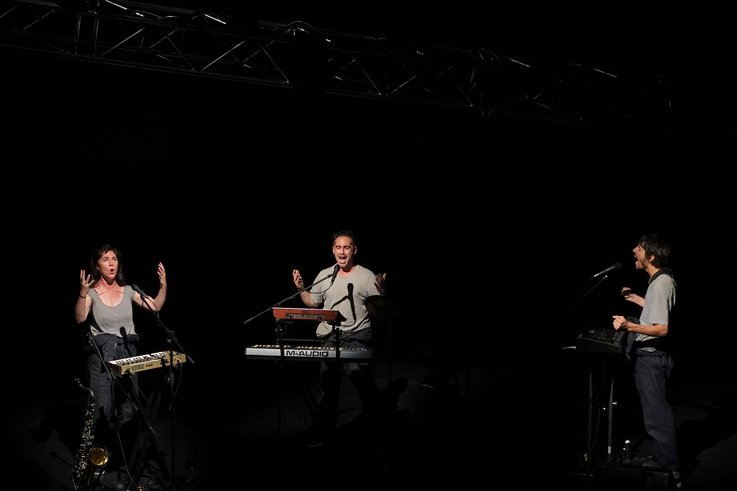 Drei Personen stehen an Mikrofonen und singen mit erhobenen Händen vor Musikinstrumenten.