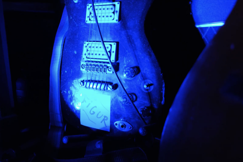 A close-up of an electric guitar in black light. A sticker with the word Figur is stuck on the guitar.
