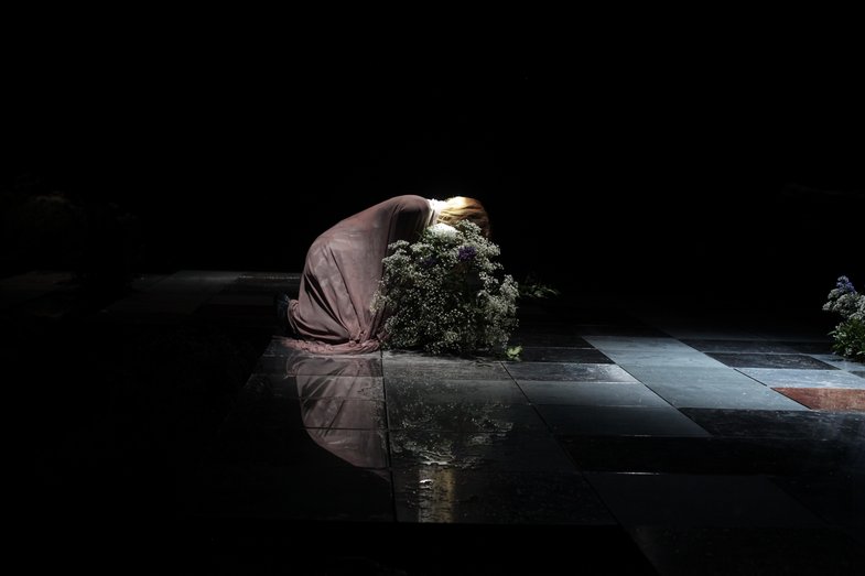 A person with blond hair kneels and presses their face into a bouquet of flowers.