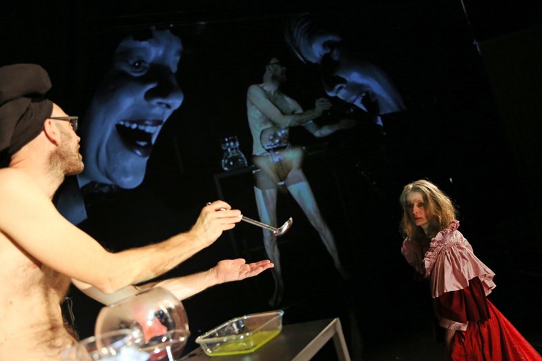 In the foreground are two people. In the background, the stage action is projected in a different perspective.