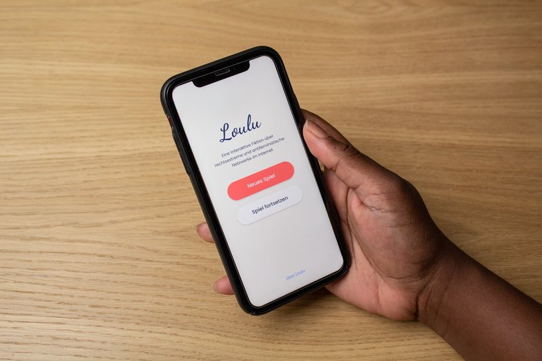 Foto einer rechten Hand, die ein Smartphone hält. Auf dem Display ist das Startbild der App „Loulu“ zu sehen. Die Hand liegt auf einem Holztisch auf. 