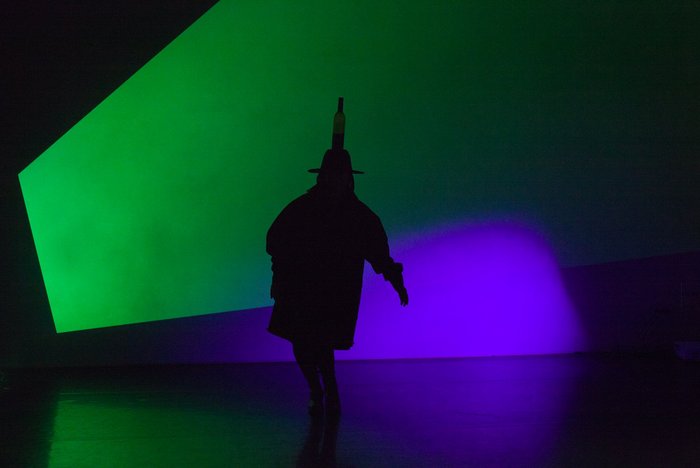 Auf einer Bühne ist der Schatten einer Figur zu sehen, die vor grüner und blauer Beleuchtung einen Hut mit einer Flasche auf dem Kopf trägt.
