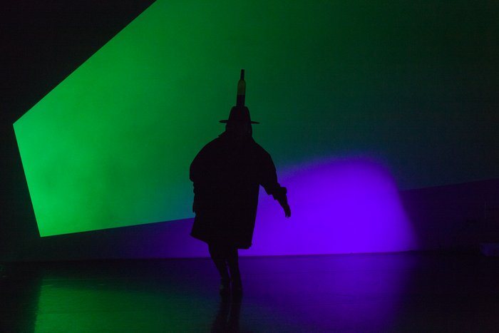 On a stage, there is a shadow of a figure in front of green and blue lighting wearing a hat with a bottle on their head.
