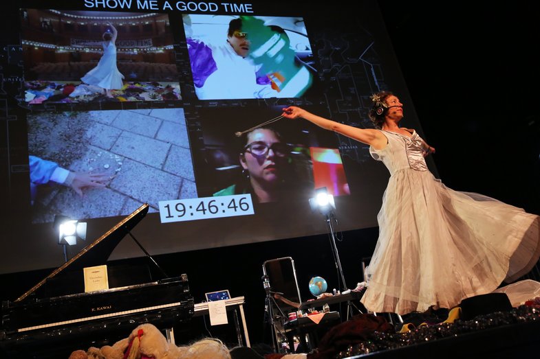 A woman in a dress stands in front of a screen with four different videos and the caption "Show me a good time".