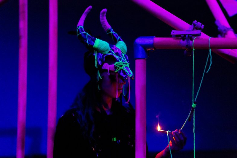A person sits in the middle of a scaffolding and holds a lit match in his hand. She wears a horned mask.