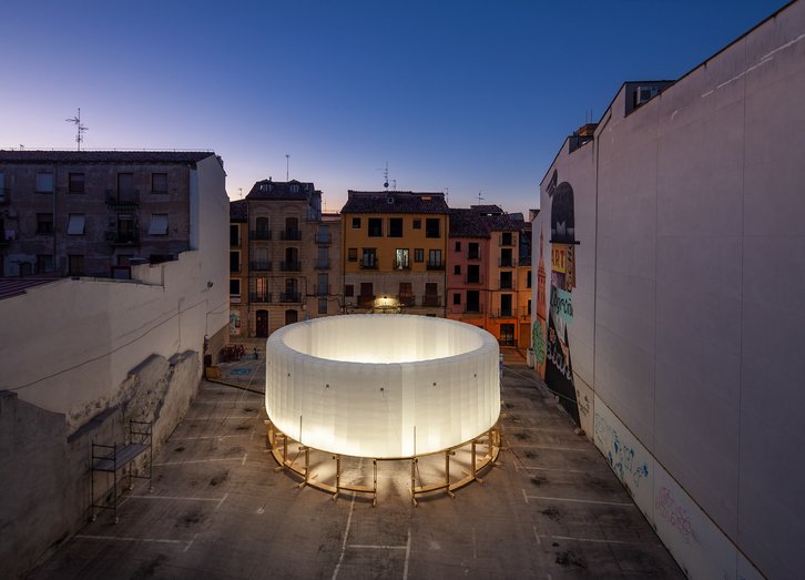 Photo of an empty parking space at dusk. A circular pavilion stands on the square. The base of the structure is a fine wooden construction, on top of which there is an inflatable structure made of white translucent textile. It is lit from within.