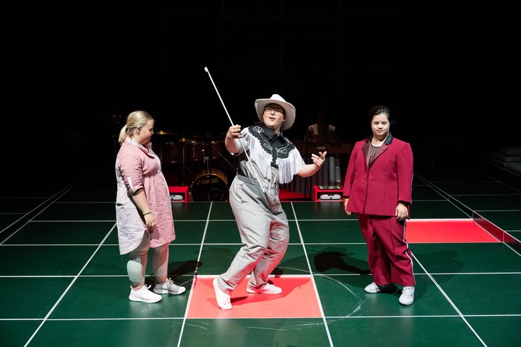 A person twirls a flag in a flared square. This person is wearing replicas of a cowboy costume. To the left and right of her are two other people watching and commenting on the performance.