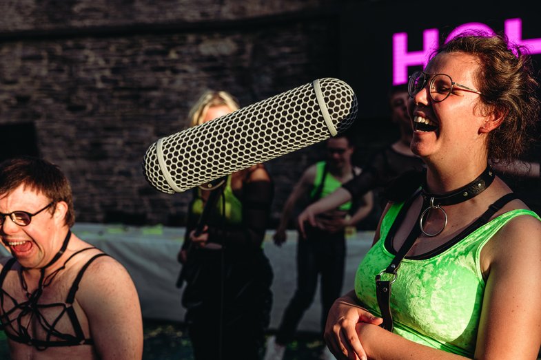 Several people dance exuberantly. One person holds a microphone boom in the face of another.