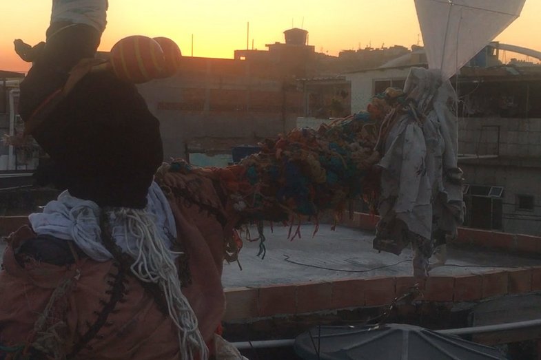 In the foreground, a person covered with cloth can be seen out of focus. This person is on the roof of a house. The sun is setting in the background.