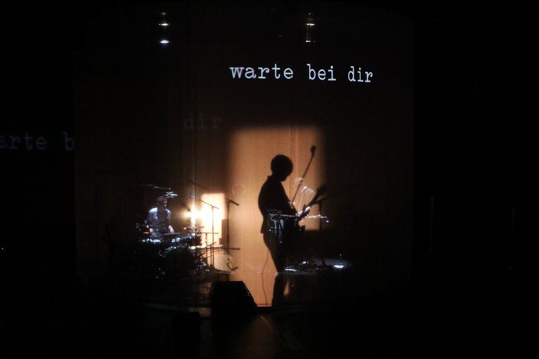 Atmosphärische Fotografie des Schattenwurfs der Gitarristin auf dem Gazestoff. Darüber sind die Worte „warte bei dir“ projiziert. 