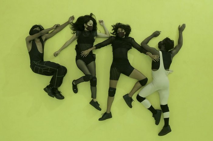 Four people lie on the floor next to each other all wearing black face masks. 