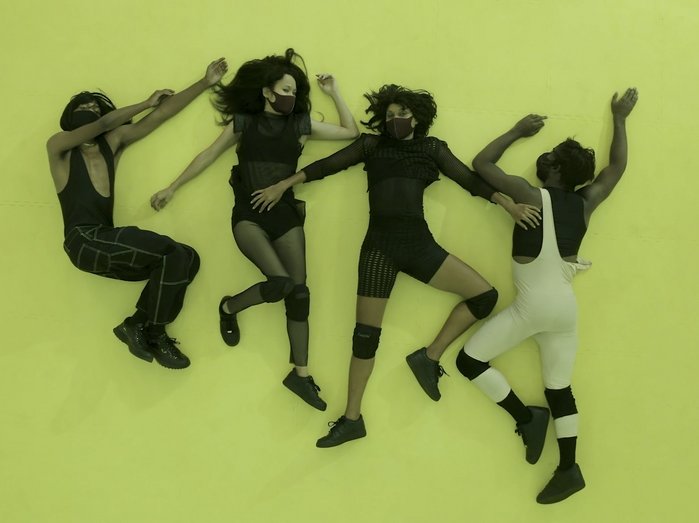 Four people lie on the floor next to each other all wearing black face masks. 