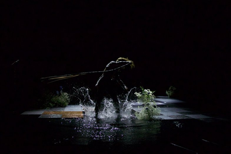 On a stage, a person stands in a puddle. The person twirls around with a long whip.