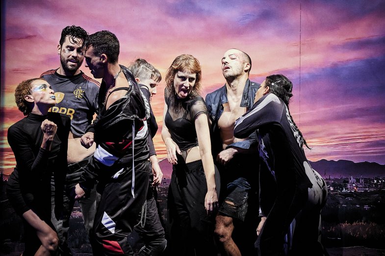 Fotografie einer Gruppe von sieben Personen in sportlicher Kleidung und Make-up. Sie sehen als, als würden sie tanzen. Im Hintergrund befindet sich eine Leinwand, auf dem ein Abendhimmel abgebildet ist.