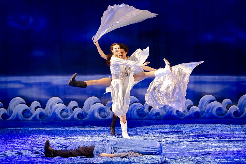 In the middle two people are dancing on the tarpaulin with silk scarves. Each person stretches one leg in the opposite direction. In front, a person is lying flat on the ground, wrapped in the tarpaulin.