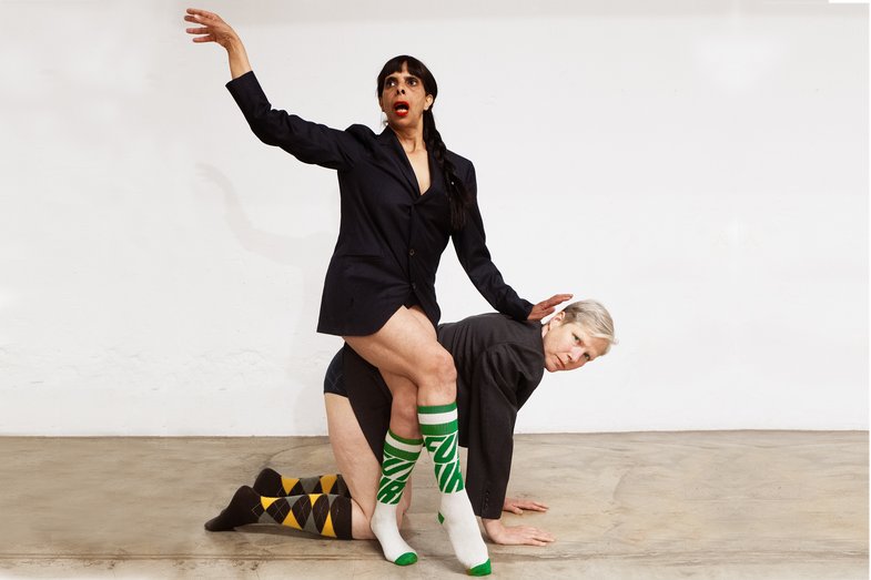 Antonia Baehr is on all fours, Latifa Laâbissi sits on Antonia's back with a lofty gesture. They are each wearing a suit jacket and knee socks.