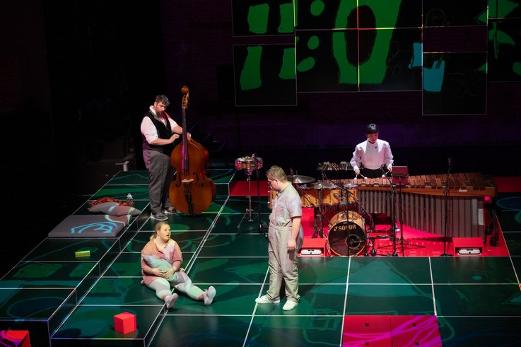 Photography of a stage situation. Two musicians accompany the scene on a double bass and percussion. One person sits on the floor with a cushion, the other person looks over at her. Some cuboids are spread out on the stage floor.