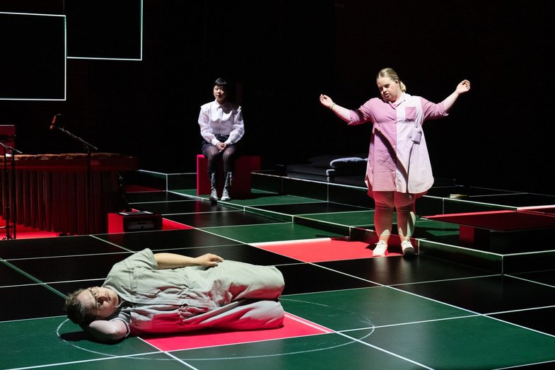 A person lies sideways in the square on display, a circle is drawn around them with chalk. A person approaches her with arms raised. In the background, another person sits on a cuboid and observes the scene.