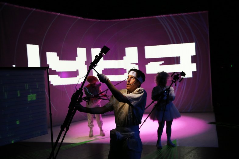 In the foreground there is a person with a technical device in their hand, perhaps a camera or a small spotlight. In the background there are two other people in front of a large picture that says "1984".