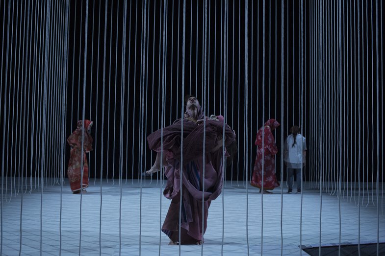 A cage of strings surrounds five people on stage. In the foreground one person carries another in their arms. The 3 others are in the background.