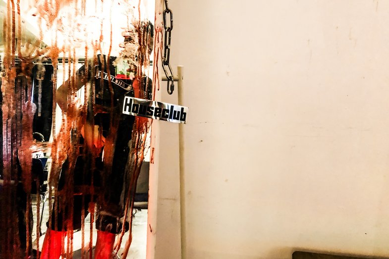 Foto aus einem Raum im Houseclub des HAU. Links im Bild ist ein Ganzkörperspiegel. Dieser ist mit roter Farbe beschmiert, das Spiegelbild zeigt unscharf eine schwarzgekleidete Person. Am Spiegel hängt rechts eine Eisenkette.