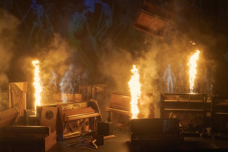 Photograph of pianos on stage, some of them erupting in fire.