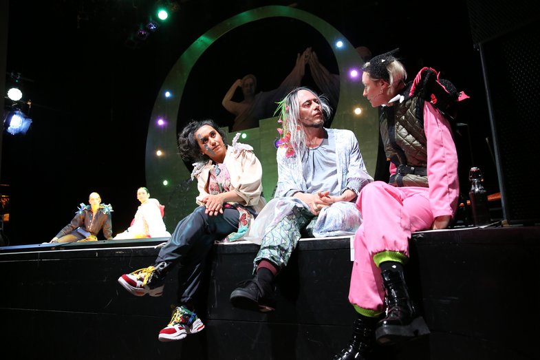 Three people sit in the foreground at the edge of the stage and talk. In the background, two people sit on the floor, behind them the ancient Greek-looking paper figures are visible.