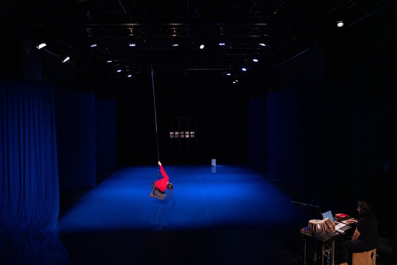 An empty stage is illuminated in blue. A white person in a red jumper holds on to a long rope and hovers above the floor.