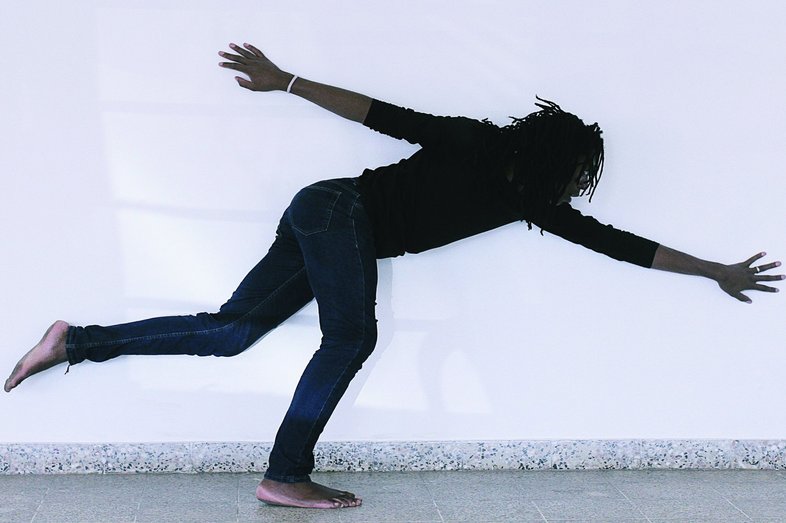 Eine Person mit Dreadlocks steht auf einem Bein, beugt sich nach vorne und streckt die Arme aus.