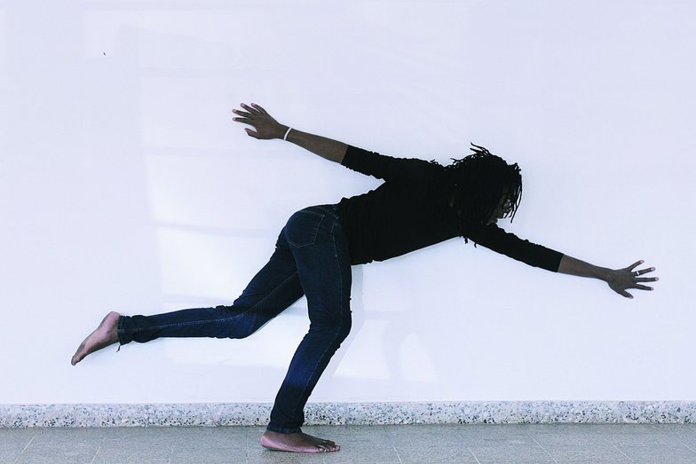 A person with dreadlocks stands on one leg, bends forward and stretches out their arms.