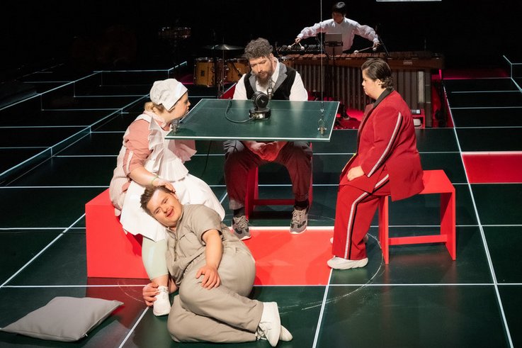 Photography of a stage scene. The stage floor consists of square panels with a board coating. One of the panels is lifted by pulleys and functions as a table. On top of it is a microphone. Three people sit around the plate, another lies at the feet of the person on the left. In the background, a person plays the xylophone.