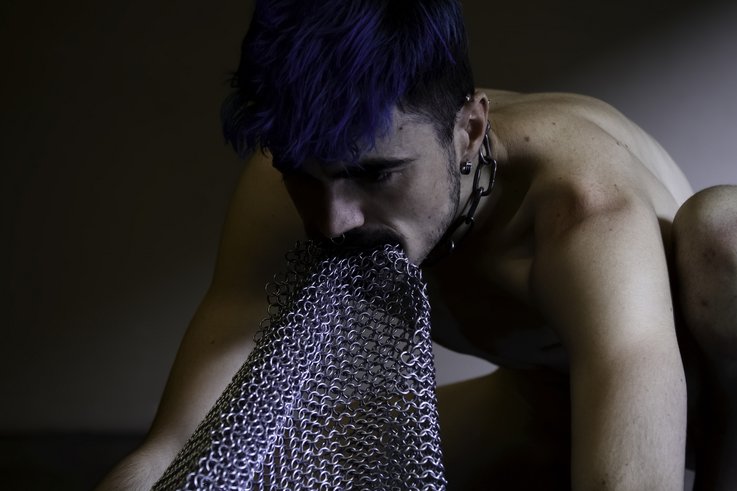 Eine Person mit nacktem Oberkörper und blauem Haar mit einem Kettenhemd, das aus ihrem Mund kommt